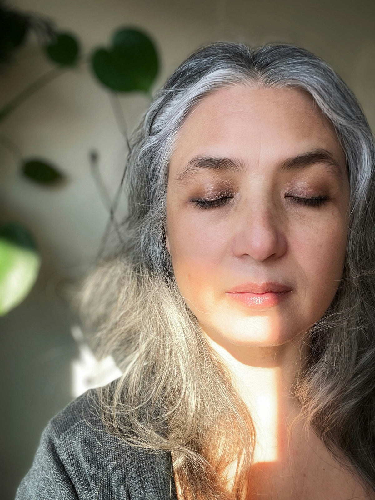 Woman in her fifties with long grey hair and eyes closed in soft window light
