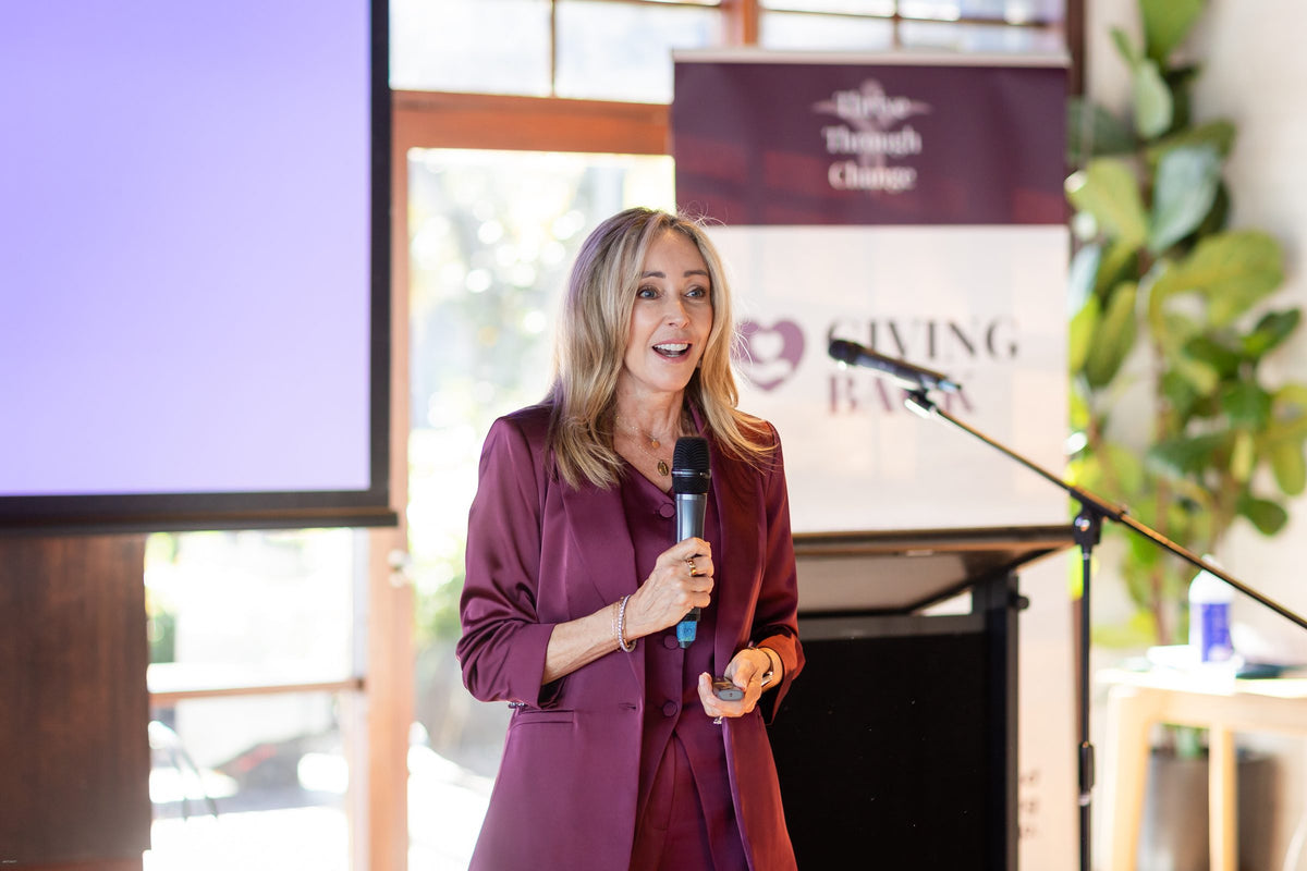 A speaker in a plum suit presenting beside the Giving Back banner at Thrive Through Change 2026